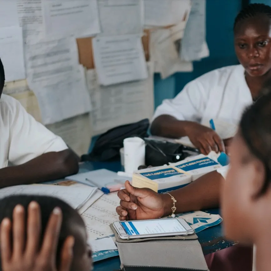 Health workers at a clinic table with patient registers, medicines and a phone, in front of a wall of records